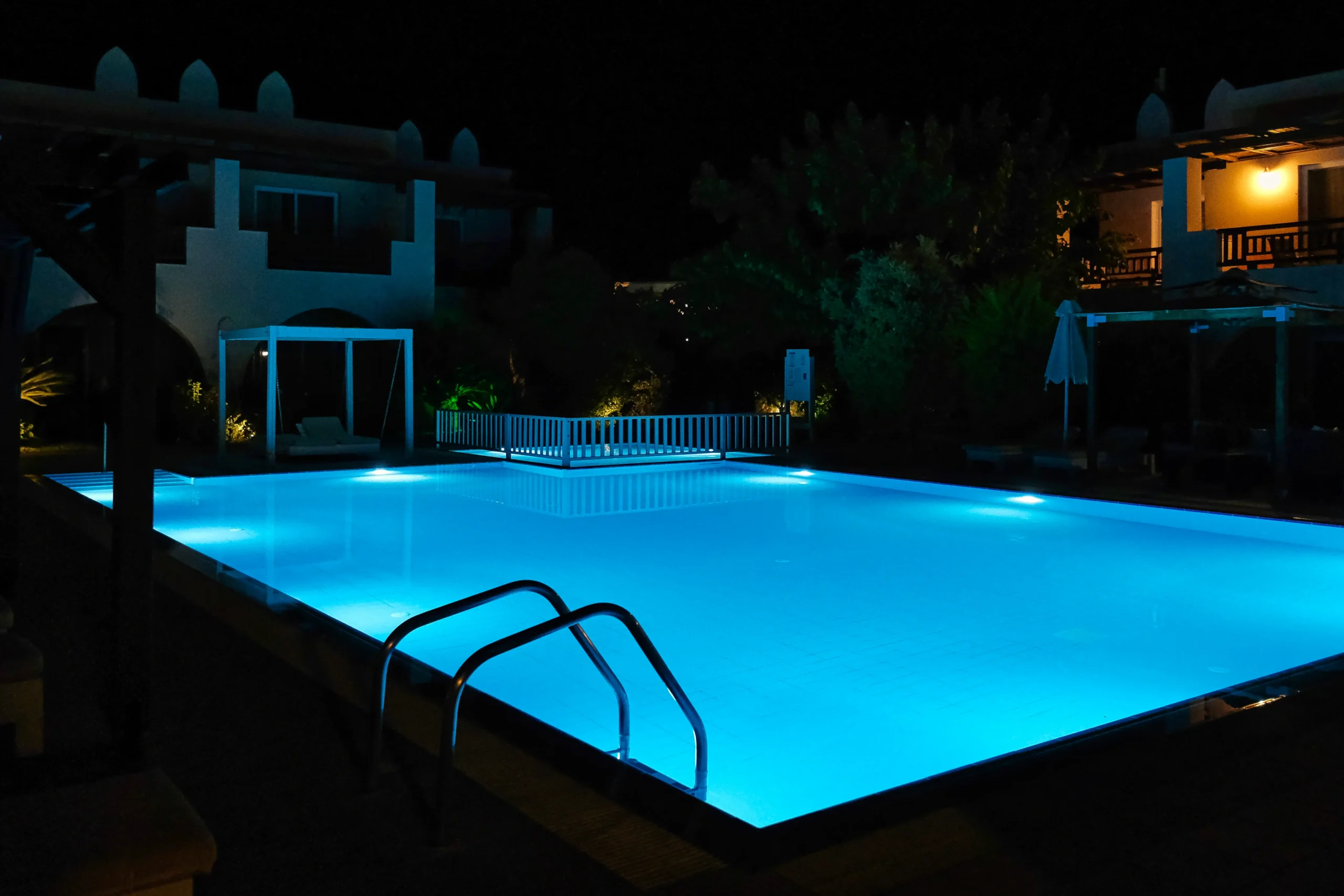 Piscina iluminada em azul à noite em área externa de hotel ou resort, com escada metálica, iluminação subaquática e arquitetura ao redor.