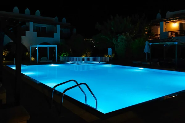 Piscina iluminada em azul à noite em área externa de hotel ou resort, com escada metálica, iluminação subaquática e arquitetura ao redor.